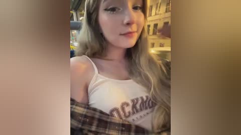 Media: Video of a young woman with long blonde hair, wearing a white Hooters tank top and plaid shirt, smiling softly, indoors.