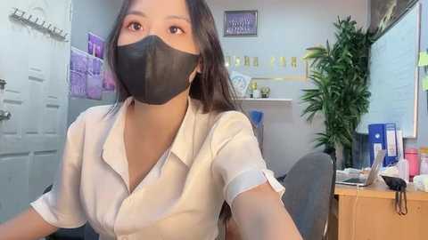 Media: Video of an East Asian woman in a white blouse, black mask, and black hair, sitting in an office with a desk, plants, and papers.