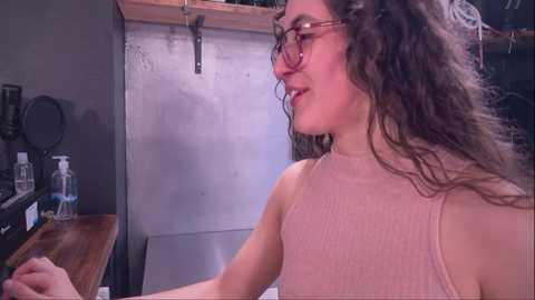 Media: Video of a woman with curly brown hair and glasses, wearing a ribbed pink tank top, sitting at a wooden table in a dimly lit, industrial-style room with metallic accents and a mounted circular mirror.