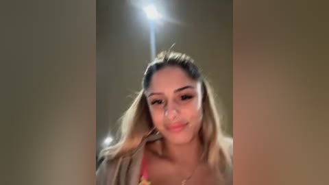 Media: Video of a young woman with light brown skin, straightened blonde hair, and a slight smile, wearing a red top, standing in a dimly lit hallway.