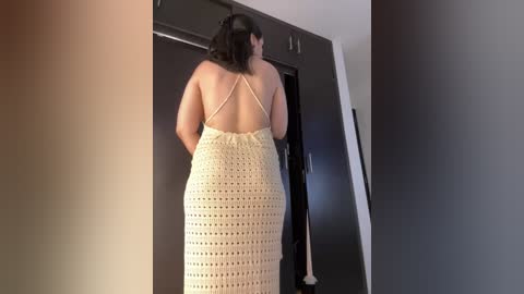 Media: Video of a woman with medium skin tone, wearing a beige crochet dress, standing in a modern room with dark wood and white walls.