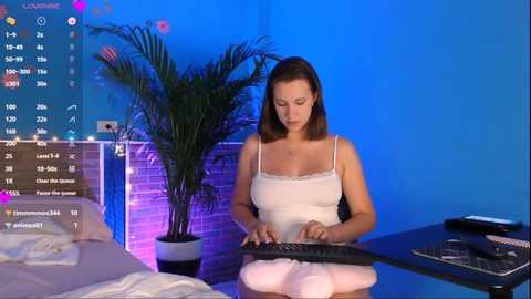 Media: A video of a light-skinned woman with straight brown hair, wearing a white camisole, typing on a computer in a modern, blue-lit room with a plant and digital screens in the background.