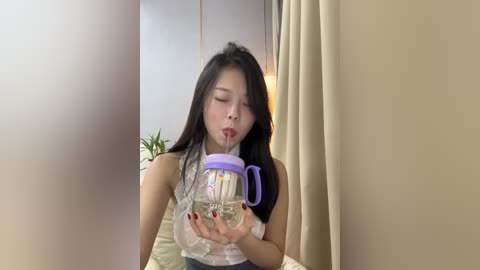 Media: Video of an Asian woman with long black hair, wearing a white lace top, drinking from a purple and clear sippy cup, in a modern, beige room with a green plant in the background.