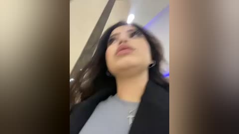 Media: A video of a woman with dark hair, wearing a black jacket over a gray shirt, looking upwards. The background is blurry, suggesting an indoor setting.