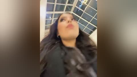 Media: A blurred video of a woman with long black hair and fair skin, wearing a black fur coat, standing indoors with a glass ceiling and grid pattern in the background.