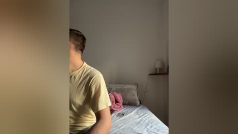 Media: Video of a young man with short hair, wearing a yellow T-shirt, sitting on a bed with a pink blanket, in a minimalist, dimly lit room with white walls and a small shelf.