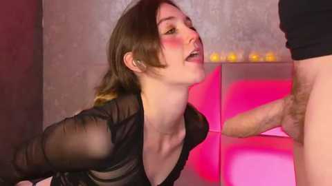 Media: Video of a Caucasian woman with fair skin, brown hair, and medium breasts, wearing a black sheer top, kneeling and licking a man's erect penis. Background features a modern, dimly lit room with pink and purple lighting.