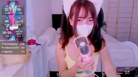 Media: Video of an Asian woman with long brown hair and a white cat headband, wearing a yellow tank top and a face mask, holding a pink hair dryer. Background shows a white room with a bed, a TV, and a fan.