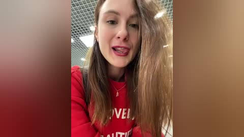 Media: Video of a young woman with long brown hair, fair skin, and red lipstick, wearing a red sweater with \"Avenue\" text. She has a slight smile and is indoors with a grid ceiling and red walls.