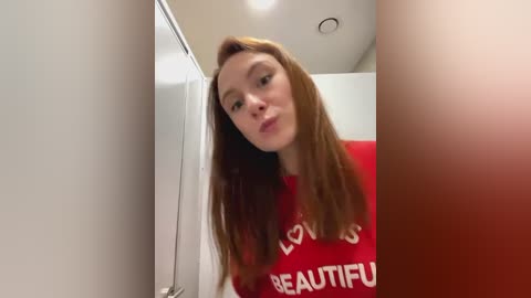 Media: Video of a fair-skinned woman with long auburn hair, wearing a red t-shirt with white text, taken in a narrow, dimly-lit bathroom with white walls.