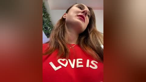 Media: A video of a woman with long brown hair wearing a red \"LOVE IS\" t-shirt, looking upward in a bright, indoor setting.