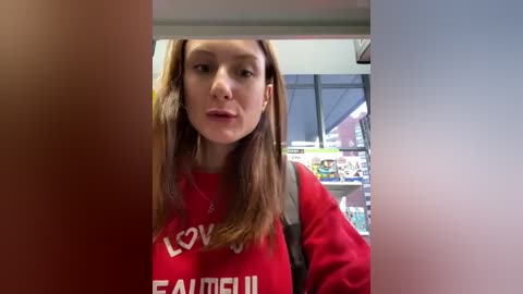 Media: Video of a young Caucasian woman with long brown hair, wearing a red sweatshirt with \"LOVE\" and \"FAMILY\" text, looking into a store. Background includes shelves with products.