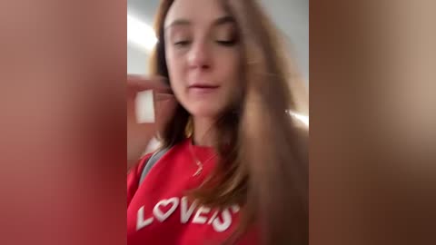 Media: Video of a young Caucasian woman with light skin, long brown hair, wearing a red \"LOVE\" sweatshirt, smiling and touching her hair, blurred background.