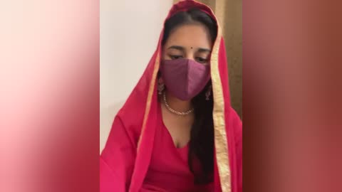 Media: Video of a South Asian woman with medium skin tone, wearing a red sari with gold trim, purple face mask, and traditional jewelry, set against a blurred background.