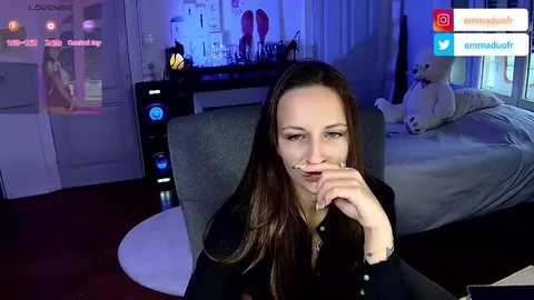 Media: Video of a young woman with long brown hair, wearing a black shirt, sitting at a desk in a dimly lit room with blue ambient lighting.