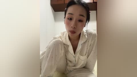 Media: Video of an Asian woman with fair skin, wearing a cream-colored, button-up shirt with subtle floral patterns, leaning against a beige wall in a dimly lit room.