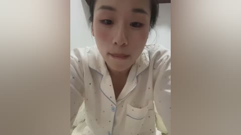 Media: A close-up video of an East Asian woman with light skin, wearing a white, patterned pajama top and looking down. Her dark hair is pulled back, and she has a neutral expression.