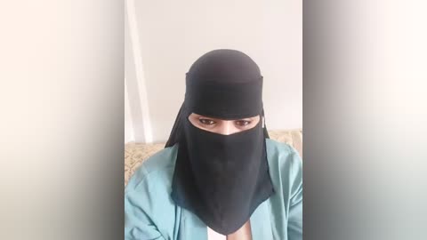 Media: Video of a woman with fair skin, wearing a black niqab that covers her face except for her eyes, and a light blue robe, sitting against a plain white wall and beige textured background.