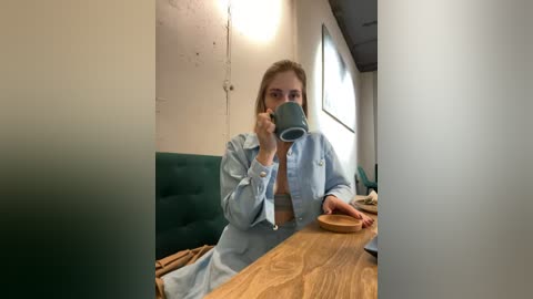 Media: A video of a woman with blonde hair, wearing a light blue shirt, drinking from a green mug at a wooden table in a cozy, dimly-lit cafe.