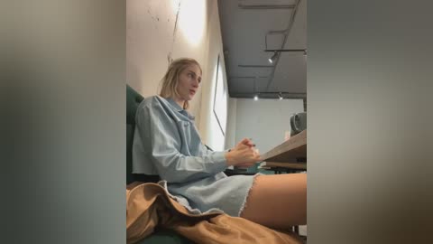 Media: Video of a young woman with fair skin, blonde hair, wearing a light blue dress, sitting on a green couch, holding a glass, in a dimly lit, industrial-style room with exposed pipes.