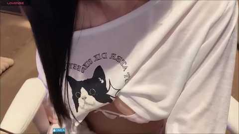 Media: A video of a woman in a white, off-the-shoulder top with a black cat graphic, leaning forward, revealing her breasts. She has long, dark hair.