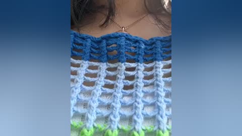 Media: Video of a woman's upper chest showing a hand-knit, multicolored (blue, white, green) crochet top with intricate patterns. The background is a smooth gradient from dark blue at the top to light blue at the bottom.