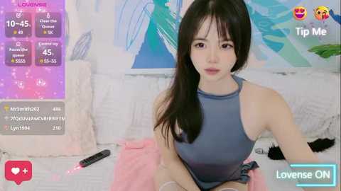 Media: A video of a young Asian woman with long black hair, fair skin, and a slender physique, wearing a sleeveless blue top, sitting on a bed with pink sheets. Background features a blue abstract painting and a TV remote.