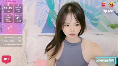 Media: Video of a young Asian woman with fair skin, long black hair, and a light gray sleeveless top. She sits on a white couch with a colorful abstract wall behind her.