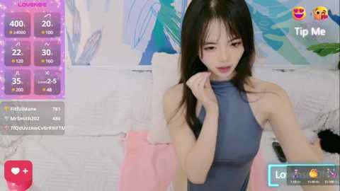 Media: A video of an Asian woman with long black hair and fair skin, wearing a blue sleeveless top, in a room with a white couch and abstract wall art.