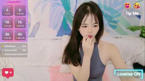 Media: Video of an East Asian woman with long black hair, wearing a sleeveless gray dress, sitting on a bed with a colorful, abstract wall painting behind her. She's biting her finger, in a casual, intimate setting.