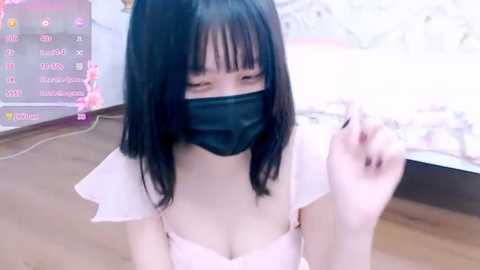 Media: A video of a young Asian woman with straight black hair and a white top, wearing a black mask, seated indoors with a bed and TV in the background.