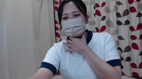 Media: Video of an Asian woman with light skin, dark hair tied back, wearing a white T-shirt and a face mask, holding her throat, indoors with floral-patterned curtains.