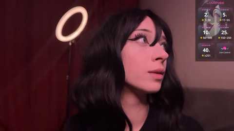 Media: Video of a young woman with fair skin, black hair, and dark-rimmed glasses, standing in a dimly lit room with a glowing halo light on the left, a digital display showing temperature and time on the right.