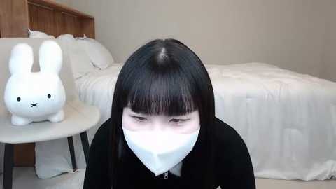 Media: Video of a young woman with straight black hair and a white face mask in a minimalist, modern bedroom. She's wearing a black sweater. The background features a white bed with fluffy pillows, a wooden headboard, and a white round side table with a bunny-shaped pillow.