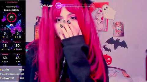 Media: Video of a young woman with long, vibrant red hair, wearing a black jacket, covering her face with her hand. The background features a bedroom with various anime posters and a cat toy.