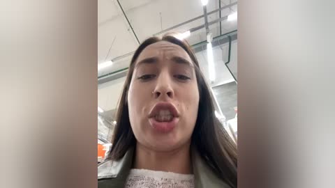 Media: Video of a woman with long brown hair, wearing a green jacket and white top, sticking her tongue out, in a warehouse with exposed beams and pipes.