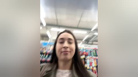 Media: A video of a young woman with long, straight black hair, fair skin, and light makeup, wearing a green jacket, standing in a brightly lit, modern supermarket. The background features colorful shelves with various products.