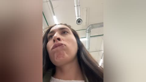 Media: Video of a young woman with long brown hair, wearing a white top, sticking her tongue out, taken from a low angle in a bright, industrial setting with visible pipes and lighting.