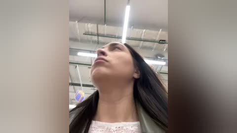 Media: A video of a young woman with long black hair, wearing a white lace top and a dark jacket, gazing upwards in a brightly lit, industrial setting with visible pipes and fluorescent lights.