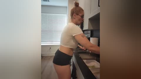 Media: Video of a curvy, light-skinned woman with blonde hair in a bun, wearing a white crop top and black shorts, bending over to open a cabinet in a modern, light-colored kitchen.