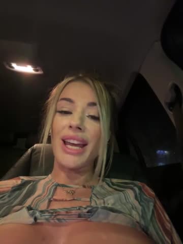 Media: Video of a smiling, blonde woman with fair skin and a pierced lip, wearing a colorful, patterned blouse, sitting in a dimly lit car.