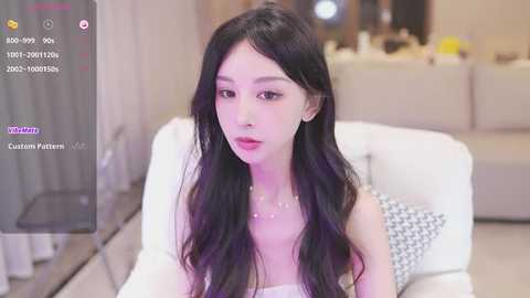 Media: Video of a fair-skinned Asian woman with long, wavy black hair, wearing a white top, seated indoors on a white couch. The background shows a modern living room with beige walls and a large window.