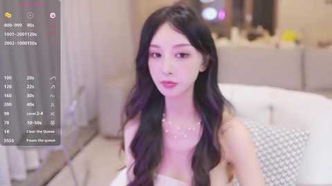 Media: Video of an East Asian woman with long, wavy black hair, wearing a white strapless dress and silver necklace, sitting on a beige couch. Background shows a blurred living room with soft lighting.