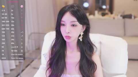 Media: Video of a fair-skinned East Asian woman with long, wavy black hair, wearing a white strapless dress and large gold earrings, seated in a modern living room with beige furnishings.