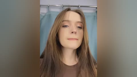 Media: A video of a young woman with long, straight, light brown hair and fair skin, wearing a brown top, standing in a bathroom with a light blue shower curtain.