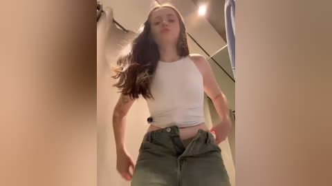 Media: Video of a slender young woman with long brown hair, wearing a white crop top and olive green high-waisted pants, standing in a dimly lit hallway.