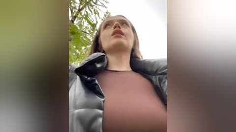 Media: Video of a young woman with light skin, straight brown hair, and a black leather jacket. She has a serious expression, looking upward. Background includes blurred green foliage.