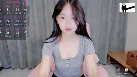 Media: A video of a young Asian woman with long black hair, wearing a grey, ribbed, low-cut top, sitting on a pink cushion. Background includes a grey curtain, wooden furniture, and a TV screen displaying a game interface.