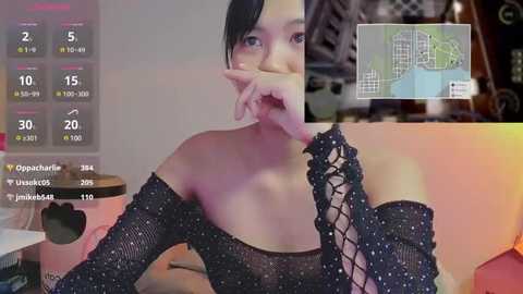 Media: A video of an Asian woman with short black hair, wearing a black, off-the-shoulder mesh top, partially covering her face with her fingers. The background features a computer screen displaying social media statistics and a map of a city.