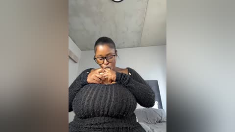 Media: A video of a pregnant Black woman with short hair, wearing black glasses and a textured sweater, sitting on a bed, holding her belly in a room with white walls and exposed concrete ceiling.